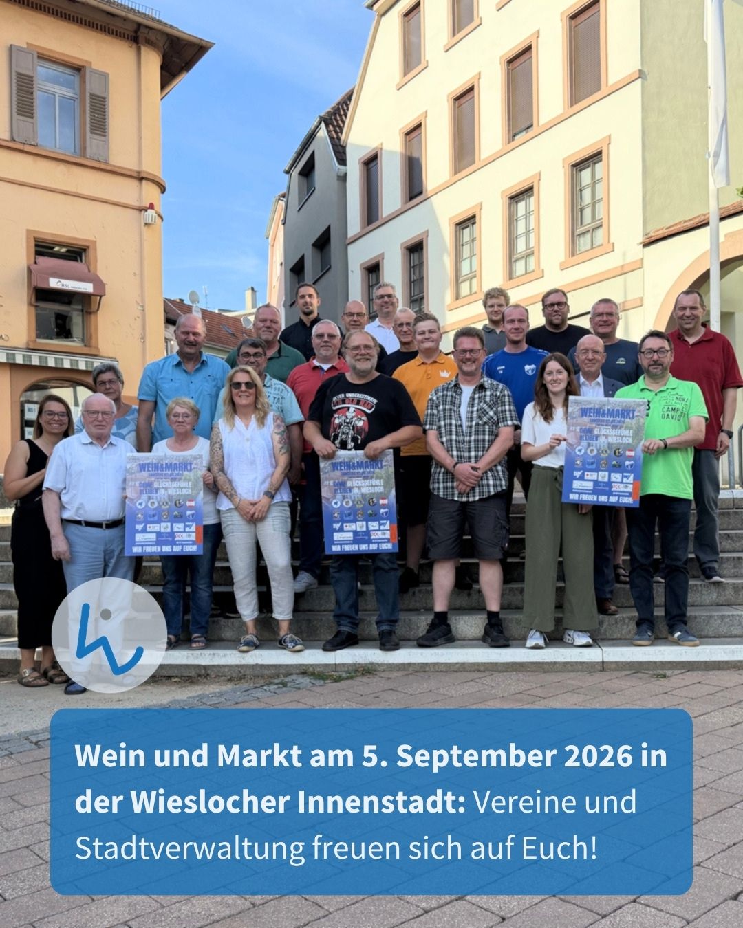 Eine große Gruppe von Personen steht auf Stufen vor Gebäuden und hält Plakate mit der Aufschrift "Wein & Markt Wiesloch". Ein blauer Textkasten unten lautet: "Wein und Markt am 5. September 2026 in der Wieslocher Innenstadt: Vereine und Stadtverwaltung freuen sich auf Euch!". (KI-Alternativtext)
