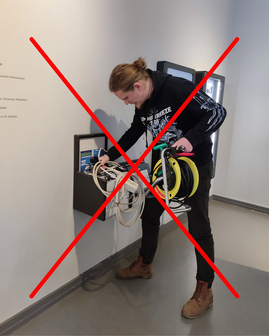 Dieser Alternativtext wurde KI-generiert: Eine Person steht vor einem Stromkasten und beschäftigt sich mit Kabeln und Geräten. Ein rotes Kreuz überdeckt einen Teil des Bildes.