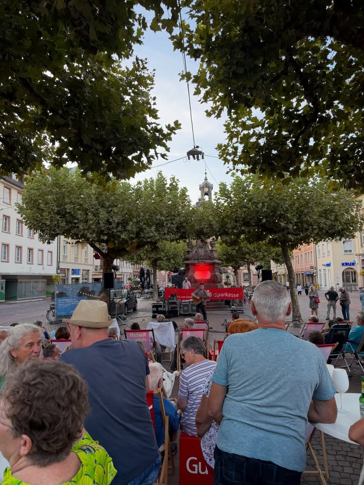Eine Gruppe von Personen sitzt auf einem Platz auf Stühlen und beobachtet eine kleine Bühne, auf der eine Person Gitarre spielt. Ein rotes Banner mit der Aufschrift "sparkasse" ist hinter dem Musiker zu sehen. Im Hintergrund erhebt sich ein Brunnen mit roter Beleuchtung zwischen Bäumen. (KI-Alternativtext)