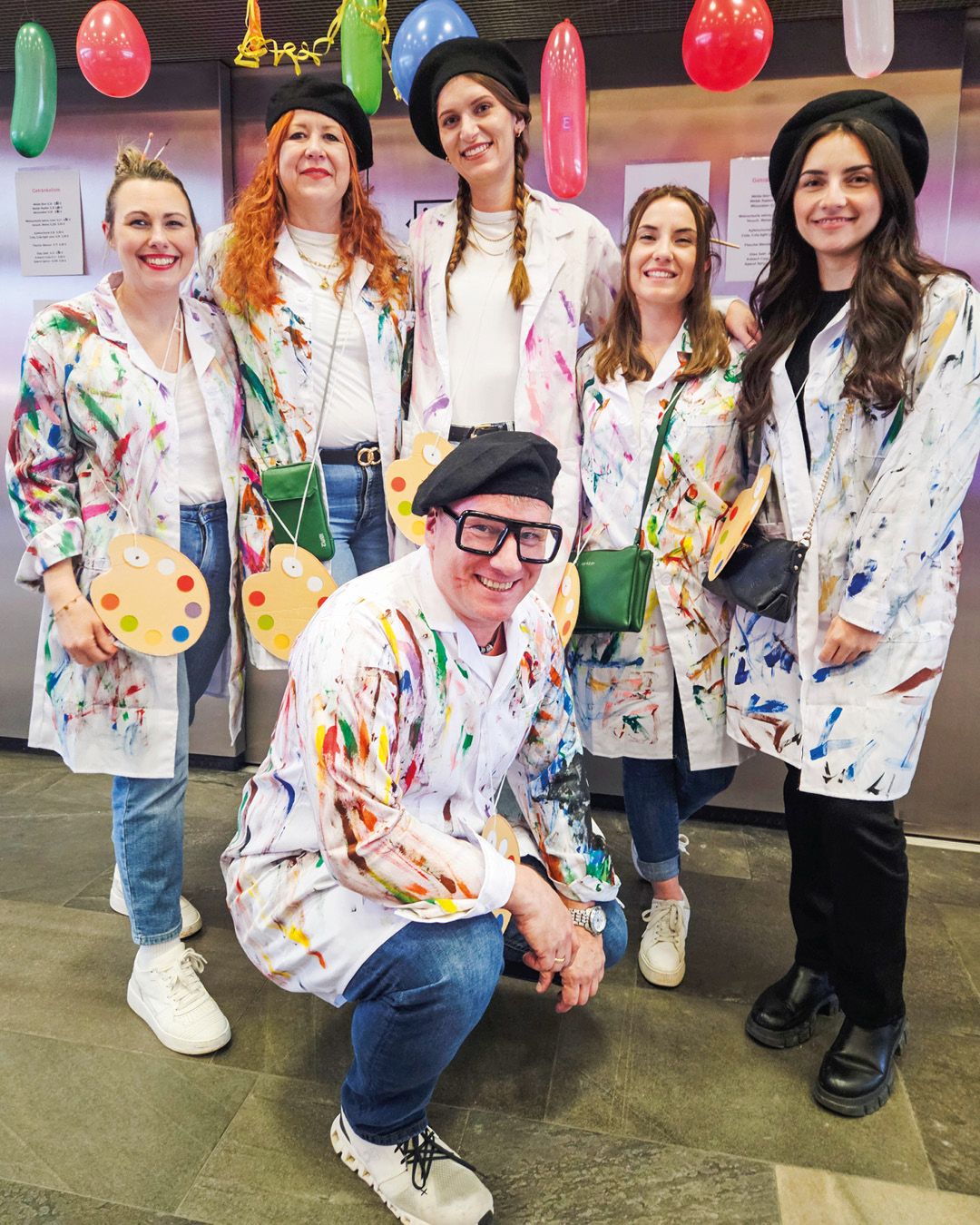 Dieser Alternativtext wurde KI-generiert: Eine Gruppe von Personen trägt farbgesprenkelte weiße Mäntel und posiert für das Foto. Einige halten Farbpaletten, und im Hintergrund sind Ballons zu sehen.