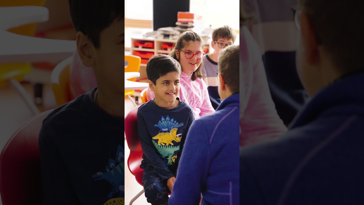 Dieser Alternativtext wurde KI-generiert: Mehrere Personen sitzen an einem Tisch, möglicherweise in einem Klassenzimmer. Sie scheinen miteinander zu interagieren und zu lächeln.