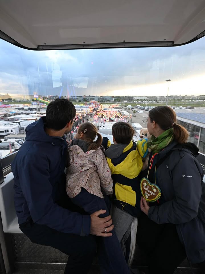 Eine Gruppe von vier Personen, darunter zwei Kinder, blickt aus einer Kabine auf einen beleuchteten Jahrmarkt mit Fahrgeschäften und Zelten. Eine Person hält ein herzförmiges Lebkuchenherz mit der Aufschrift „Frohes Fest“. (KI-Alternativtext)