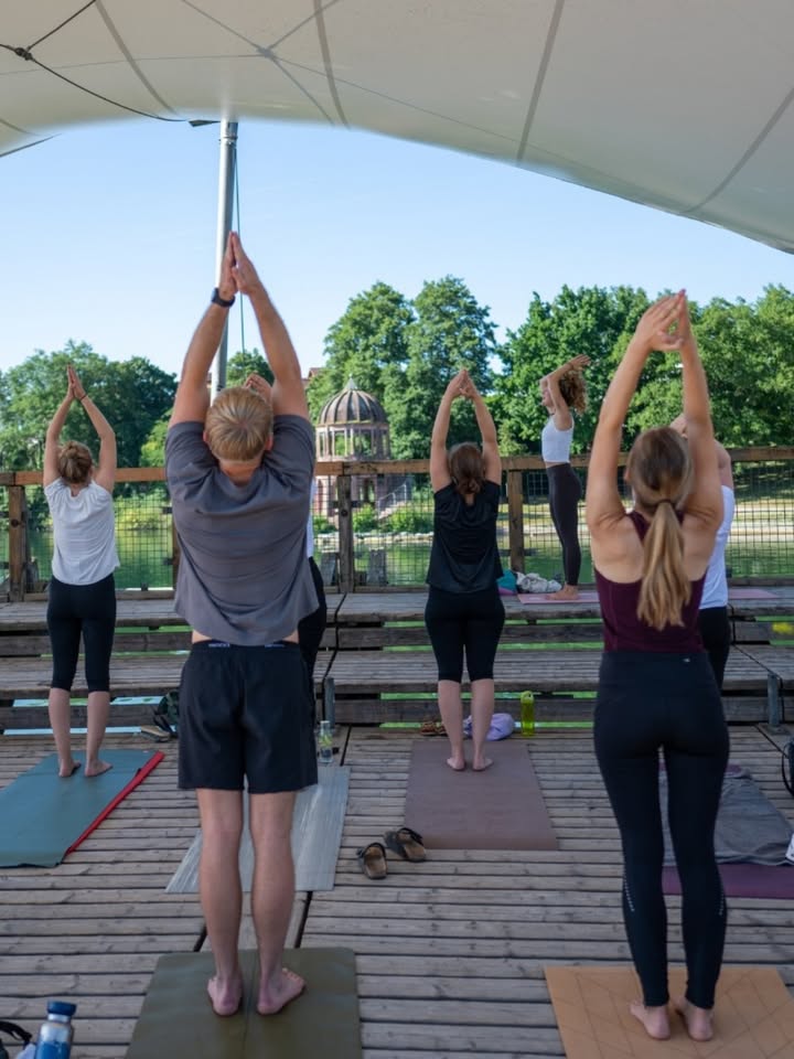 Mehrere Personen stehen auf Yogamatten unter einem Zeltdach, die Arme über dem Kopf verschränkt, und üben eine Yoga-Pose. Im Hintergrund sind Bäume, ein Gewässer und ein Pavillon zu sehen. (KI-Alternativtext)