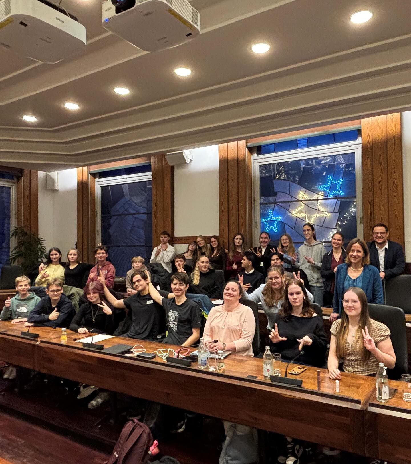 Dieser Alternativtext wurde KI-generiert: Eine Gruppe von Personen sitzt an langen Tischen in einem Raum mit Holzvertäfelung. Im Hintergrund ist ein Fenster mit Buntglas und ein Projektor an der Decke zu sehen.