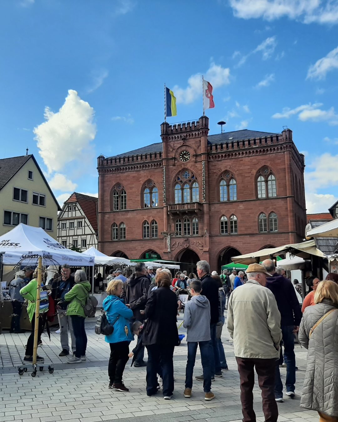 Dieser Alternativtext wurde KI-generiert: Ein historisches Gebäude mit einem markanten Turm steht im Zentrum eines belebten Marktplatzes. Menschengruppen durchstreifen den Platz, der von Marktständen beschattet wird.