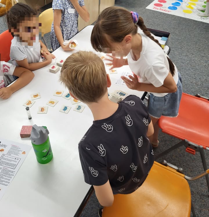 Mehrere Kinder sitzen um einen weißen Tisch und spielen ein Kartenspiel. Ein Kind im Vordergrund sitzt mit dem Rücken zur Kamera, während ein anderes Kind steht und mit den Händen gestikuliert. Auf dem Tisch liegen verstreute Spielkarten, ein Stapel Karten, eine grüne Trinkflasche und ein Blatt Papier. (KI-Alternativtext)