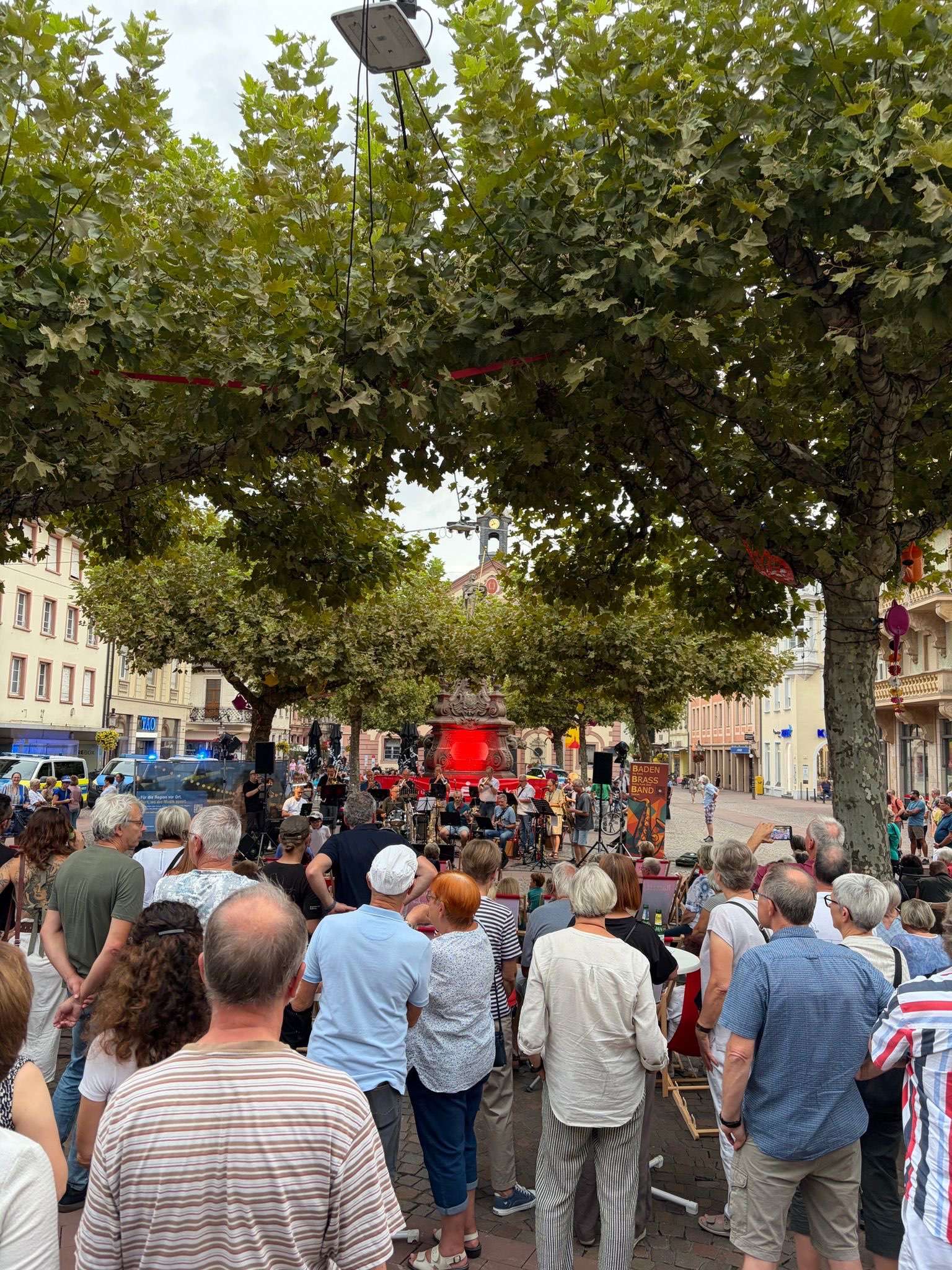 Eine Menschenmenge steht auf einem Platz unter Bäumen und schaut auf eine Bühne, auf der die Baden Brass Band spielt. Im Hintergrund leuchtet ein Brunnen rot. (KI-Alternativtext)