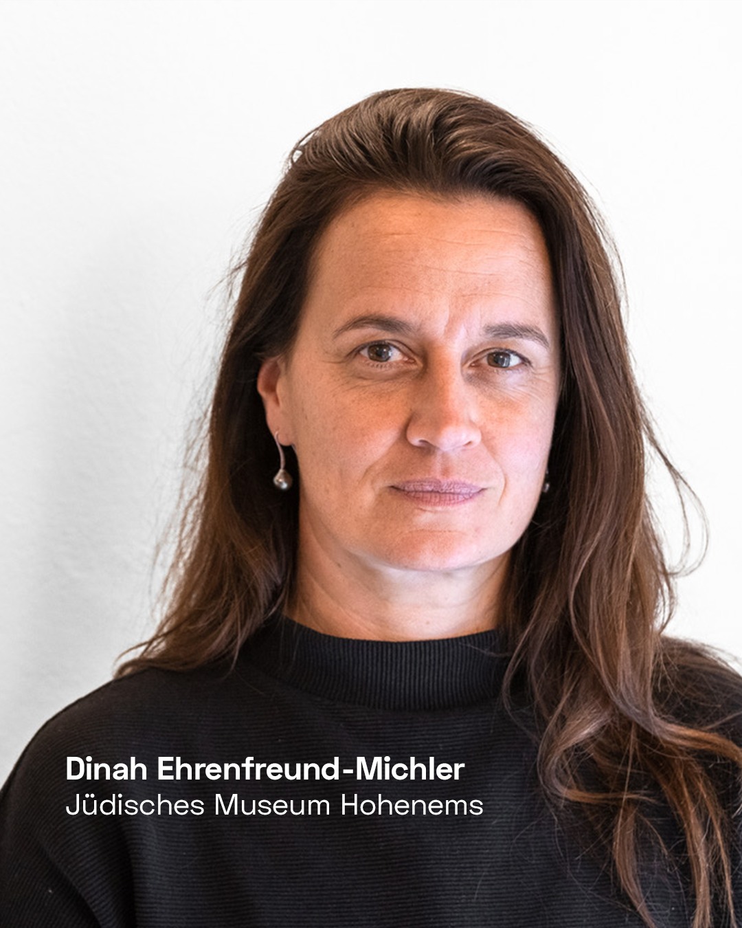 Eine Person mit langen braunen Haaren und schwarzem Rollkragenpullover blickt frontal in die Kamera. Im unteren Bildbereich steht der weiße Text: „Dinah Ehrenfreund-Michler, Jüdisches Museum Hohenems“. (KI-Alternativtext)