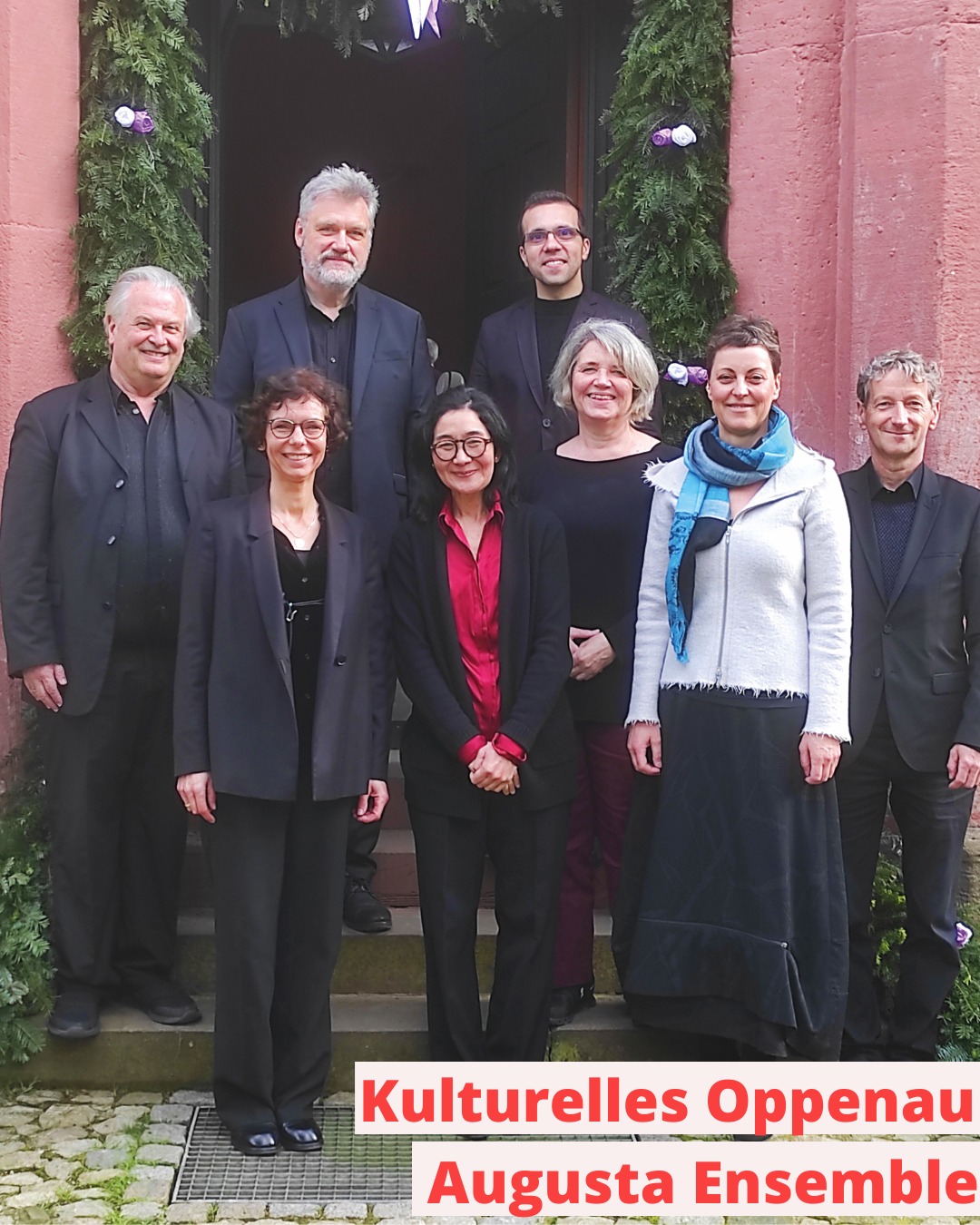 Dieser Alternativtext wurde KI-generiert: Eine Gruppe von Personen steht vor einem roten Gebäude mit Dekorationen. Ein Schild mit Text ist im Vordergrund zu sehen.