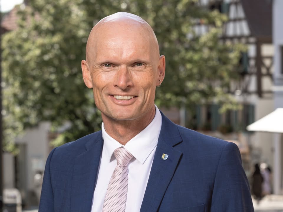 Dieser Alternativtext wurde KI-generiert: Porträt einer Person mit kurzen Haaren, die in einen Anzug gekleidet ist und lächelt. Im Hintergrund sind Gebäude und Bäume zu sehen.