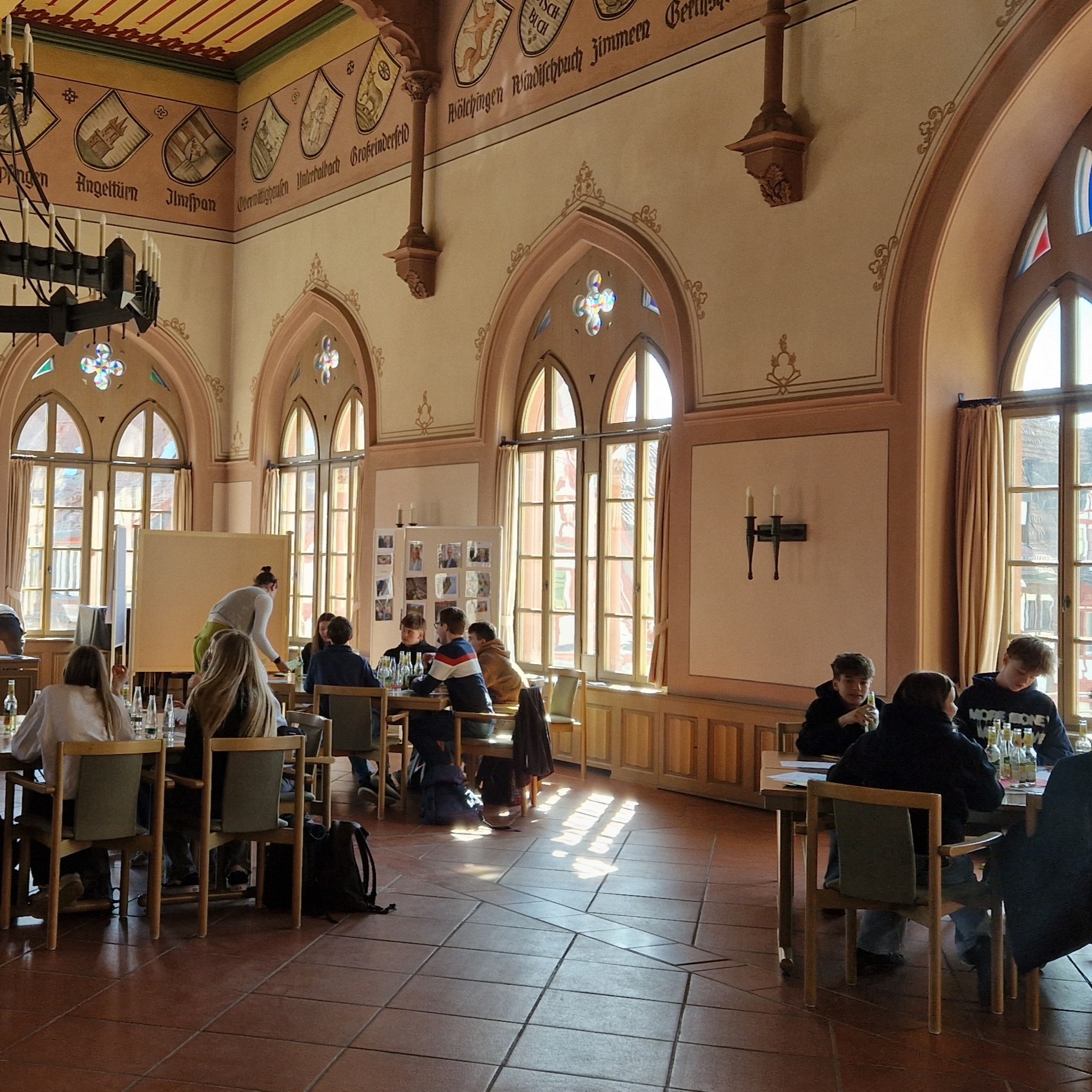 Menschen sitzen an Holztischen in einem großen, hellen Saal mit gotischen Fenstern und Wappen an den Wänden; eine Person steht vor einer Tafel und spricht mit einer Gruppe. (KI-Alternativtext)