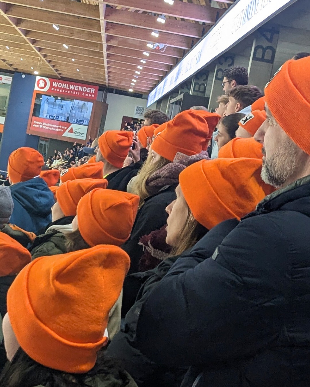 Dieser Alternativtext wurde KI-generiert: Eine Menschenmenge trägt auffällige orangefarbene Strickmützen. Im Hintergrund ist eine große Werbefläche und eine hallenartige Struktur zu sehen.
