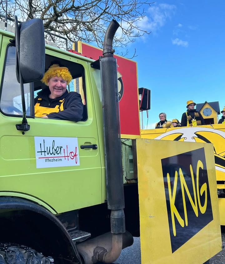 Dieser Alternativtext wurde KI-generiert: Ein grüner Truck mit einer Person am Steuer und gelbem Kopfschmuck. Im Hintergrund sind weitere Personen in ähnlicher Kleidung und ein Festwagen erkennbar.