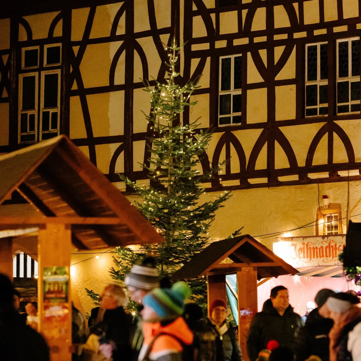 Dieser Alternativtext wurde KI-generiert: Ein festlich geschmückter Weihnachtsbaum vor einem Gebäude mit Fachwerkmuster. Menschen sind im Hintergrund zu sehen, vermutlich auf einem Weihnachtsmarkt.