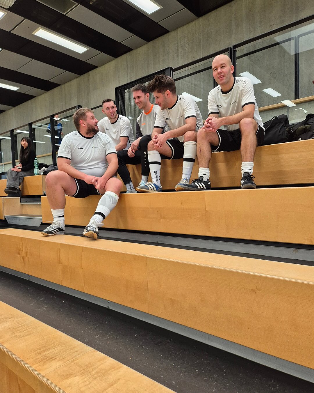 Dieser Alternativtext wurde KI-generiert: Mehrere Personen in heller Sportbekleidung sitzen auf einer hölzernen Sitzbank in einem Raum mit Spinden. Sie wirken entspannt und unterhalten sich miteinander.