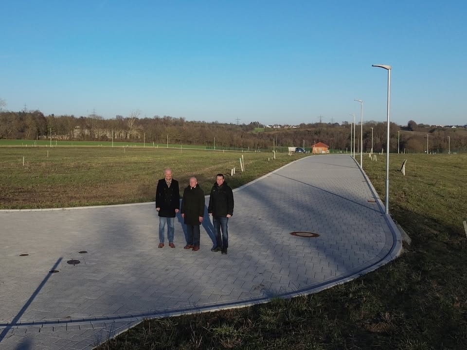 Dieser Alternativtext wurde KI-generiert: Drei Personen gehen auf einem gepflasterten Weg durch eine ländliche Landschaft. Im Hintergrund sind Bäume und ein blauer Himmel zu sehen.