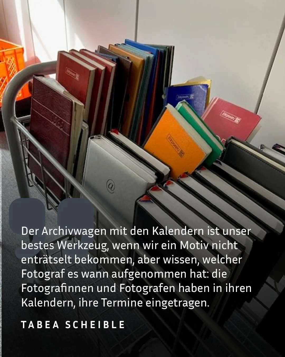 Ein Metall-Archivwagen ist mit zahlreichen bunten Kalendern gefüllt, darunter rote, blaue, orange und grüne Exemplare sowie ein weißer Kalender mit einem "@"-Symbol. Im unteren Bereich steht ein Zitat: „Der Archivwagen mit den Kalendern ist unser bestes Werkzeug... die Fotografinnen und Fotografen haben in ihren Kalendern ihre Termine eingetragen." Darunter steht der Name Tabea Scheible. (KI-Alternativtext)