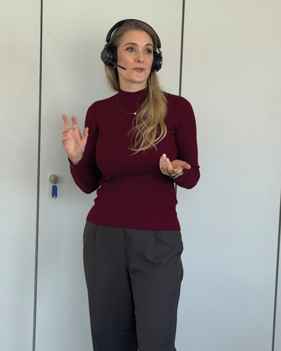 Dieser Alternativtext wurde KI-generiert: Eine Person mit Kopfhörern gestikuliert während sie spricht. Sie trägt dunkle Kleidung vor einem hellen Hintergrund.