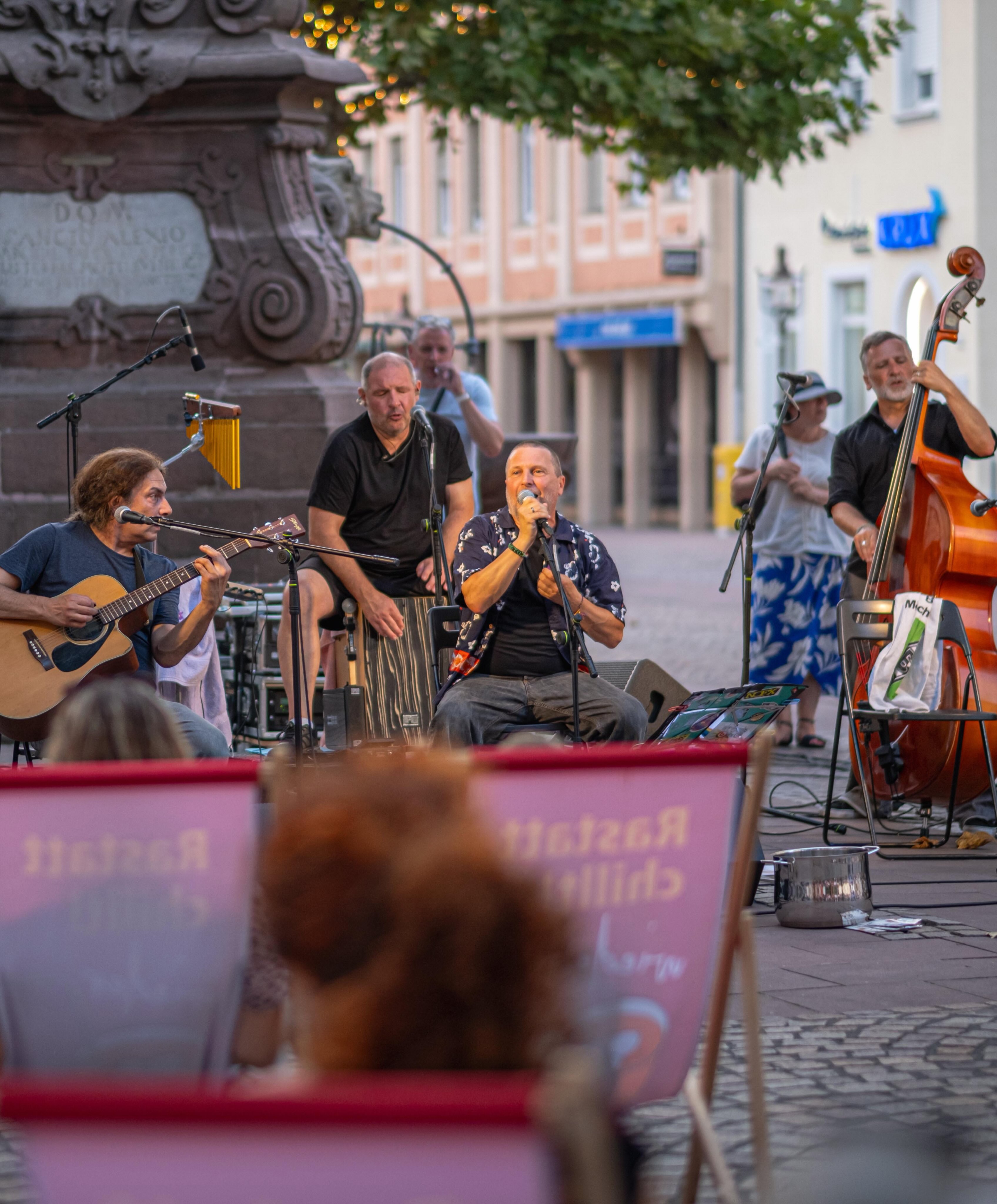 Eine Musikgruppe spielt im Freien auf einem gepflasterten Platz. Eine Person spielt Akustikgitarre, eine andere singt in ein Mikrofon, eine dritte bedient ein hohes Schlaginstrument und eine vierte spielt einen Kontrabass. Im unscharfen Vordergrund sind Köpfe von Zuhörern und ein rosa Schild zu sehen. (KI-Alternativtext)