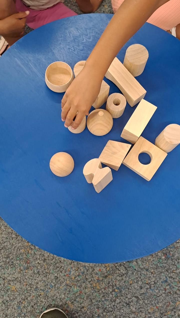 Eine Hand greift nach einem kleinen Holzbaustein auf einem blauen Tisch. Verschiedene geometrische Holzformen wie Würfel, Zylinder und Kugeln liegen verstreut darauf. (KI-Alternativtext)