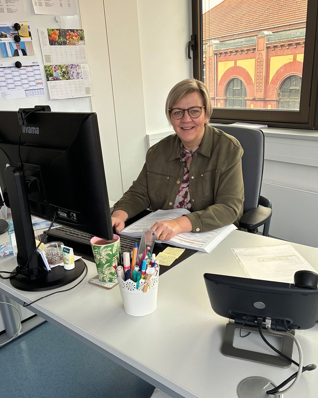 Dieser Alternativtext wurde KI-generiert: Eine Person sitzt an einem Schreibtisch mit einem Computer und Papierstapeln. Im Hintergrund ist ein Fenster mit Blick auf ein Gebäude zu sehen.