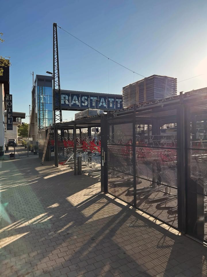 Dieser Alternativtext wurde KI-generiert: Ein Gebäude mit einem Schild darüber und einem Bereich mit Sitzgelegenheiten im Freien. Sonnenlicht wirft Schatten auf den gepflasterten Boden.