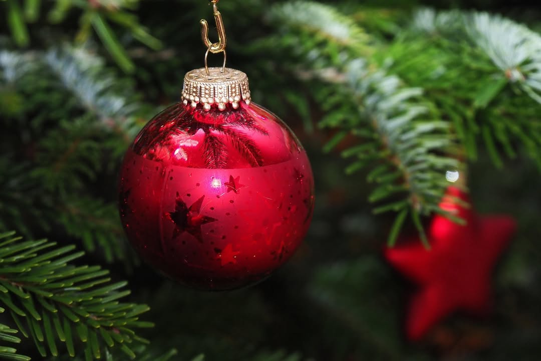 Dieser Alternativtext wurde KI-generiert: Ein roter Weihnachtsschmuck hängt an einem grünen Tannenzweig. Im Hintergrund ist ein roter Stern erkennbar.