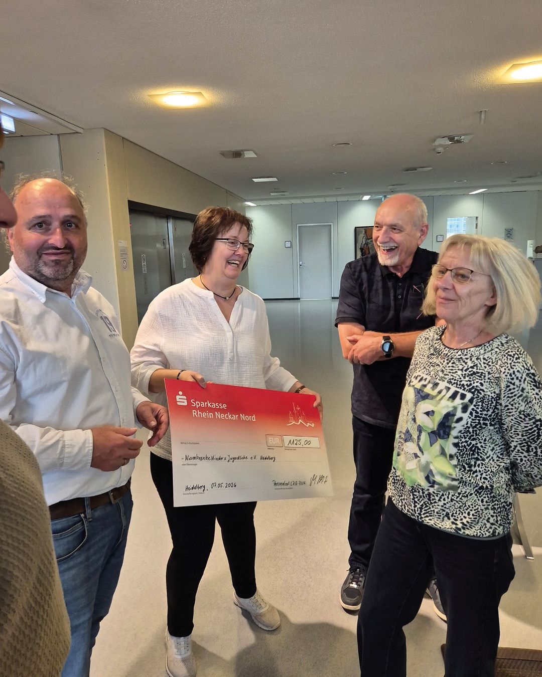 Eine Gruppe von vier Personen steht in einem Flur, eine hält einen großen Spendenscheck der Sparkasse Rhein Neckar Nord über 1.125 Euro an die Nachwuchs-Kinder- und Jugendhilfe e.V. Heidelberg vom 07.05.2024. Alle lächeln und wirken zufrieden. (KI-Alternativtext)