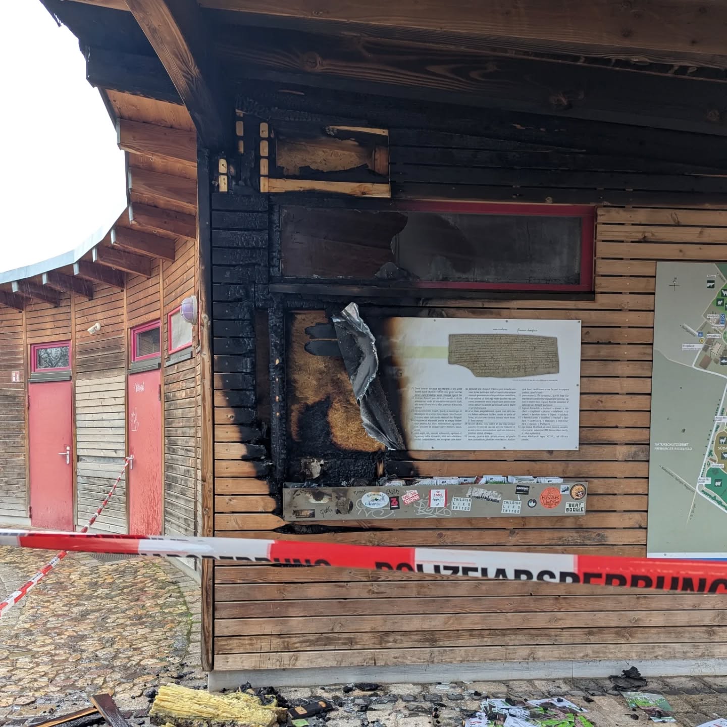 Dieser Alternativtext wurde KI-generiert: Ein Holzgebäude zeigt deutliche Brandschäden an einer Ecke. Im Vordergrund eine Absperrung mit rotem Flatterband, rechts ein Informationsbrett.