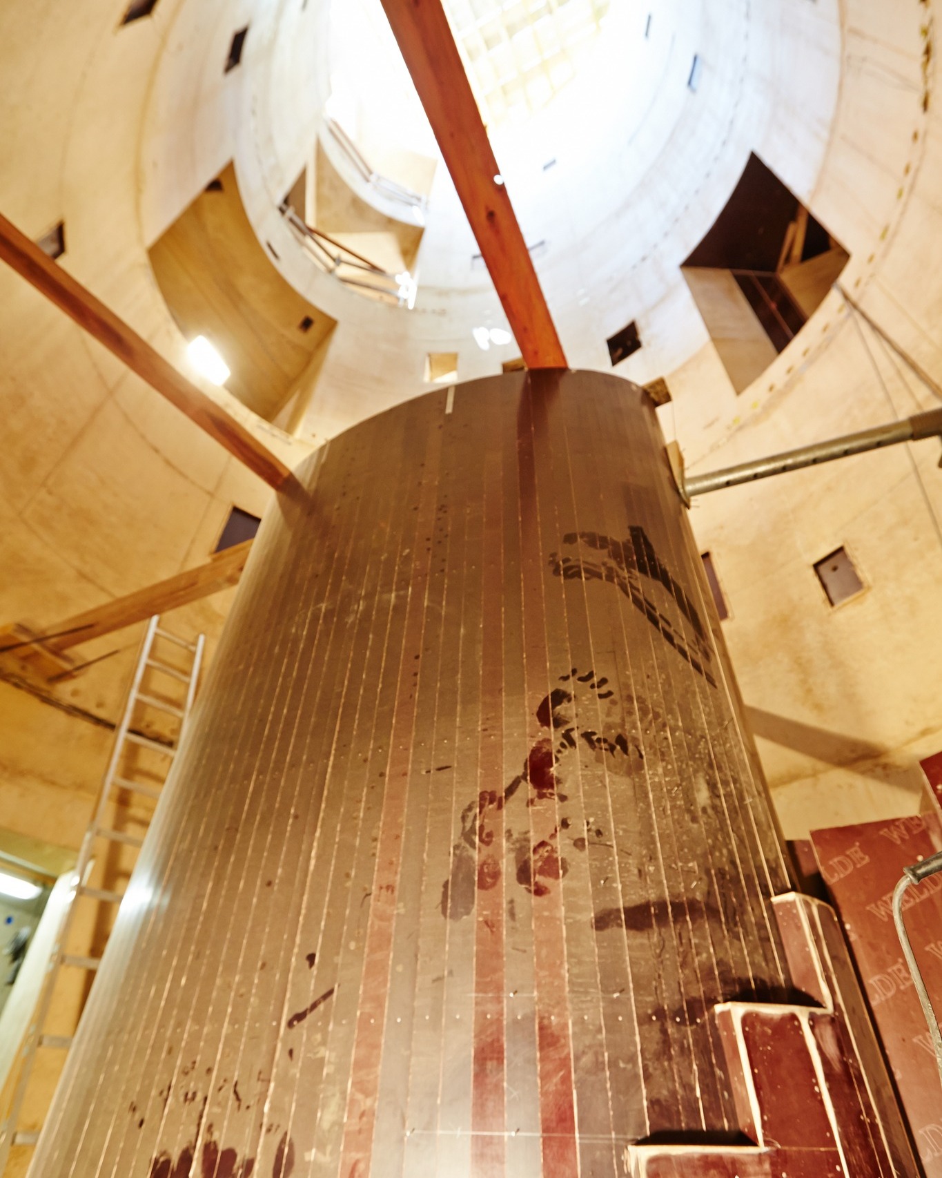 Dieser Alternativtext wurde KI-generiert: Ein Innenraum zeigt einen hohen, zylindrischen Holzbalken, der von einer hölzernen Konstruktion umgeben ist. Die Aufnahme ist von unten nach oben gerichtet.