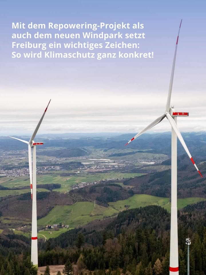 Dieser Alternativtext wurde KI-generiert: Ein farbiges Bild zeigt mehrere Windkraftanlagen vor einer hügeligen Landschaft und einer Stadt im Hintergrund. Der Himmel ist blau mit vereinzelten Wolken.
