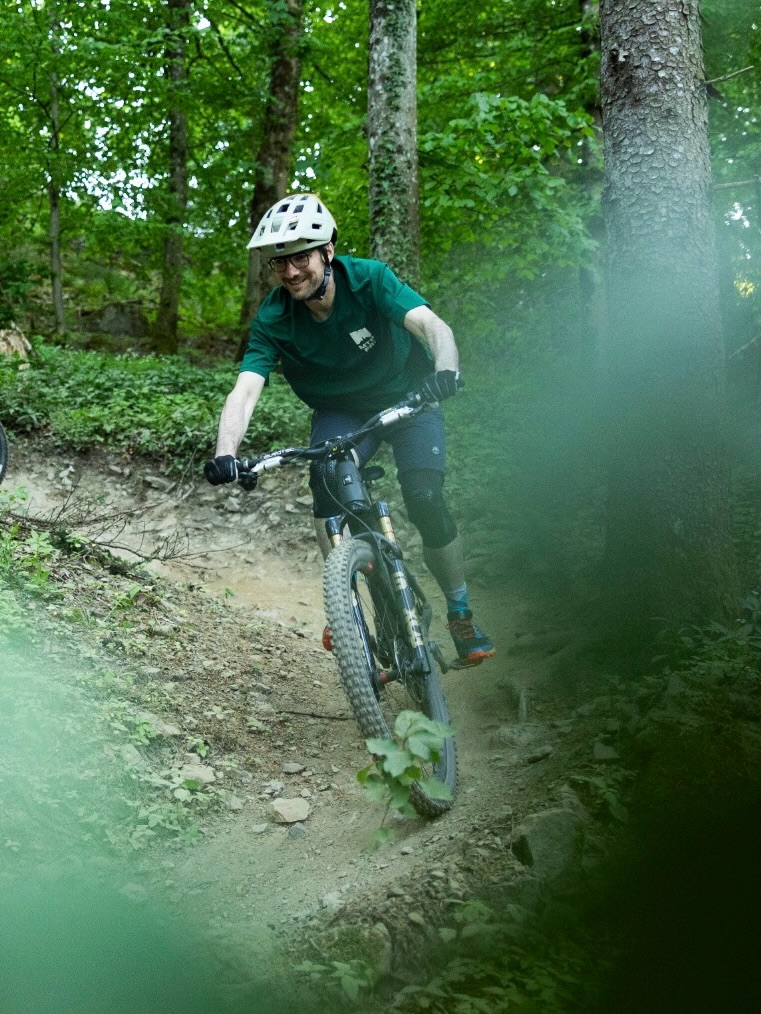 Dieser Alternativtext wurde KI-generiert: Eine Person fährt mit einem Mountainbike auf einem Waldweg. Das Bild ist farbig und zeigt eine Bewegung, die durch Unschärfe dargestellt wird.