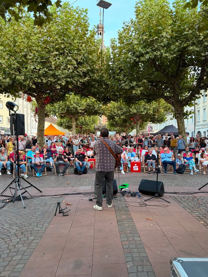 Eine Person steht mit dem Rücken zur Kamera auf einem gepflasterten Platz und spielt Gitarne. Vor der Person sitzt und steht ein großes Publikum, das die Aufführung beobachtet. Im Hintergrund rahmen große Bäume die Szene ein, dahinter ist ein Kirchturm sichtbar. (KI-Alternativtext)