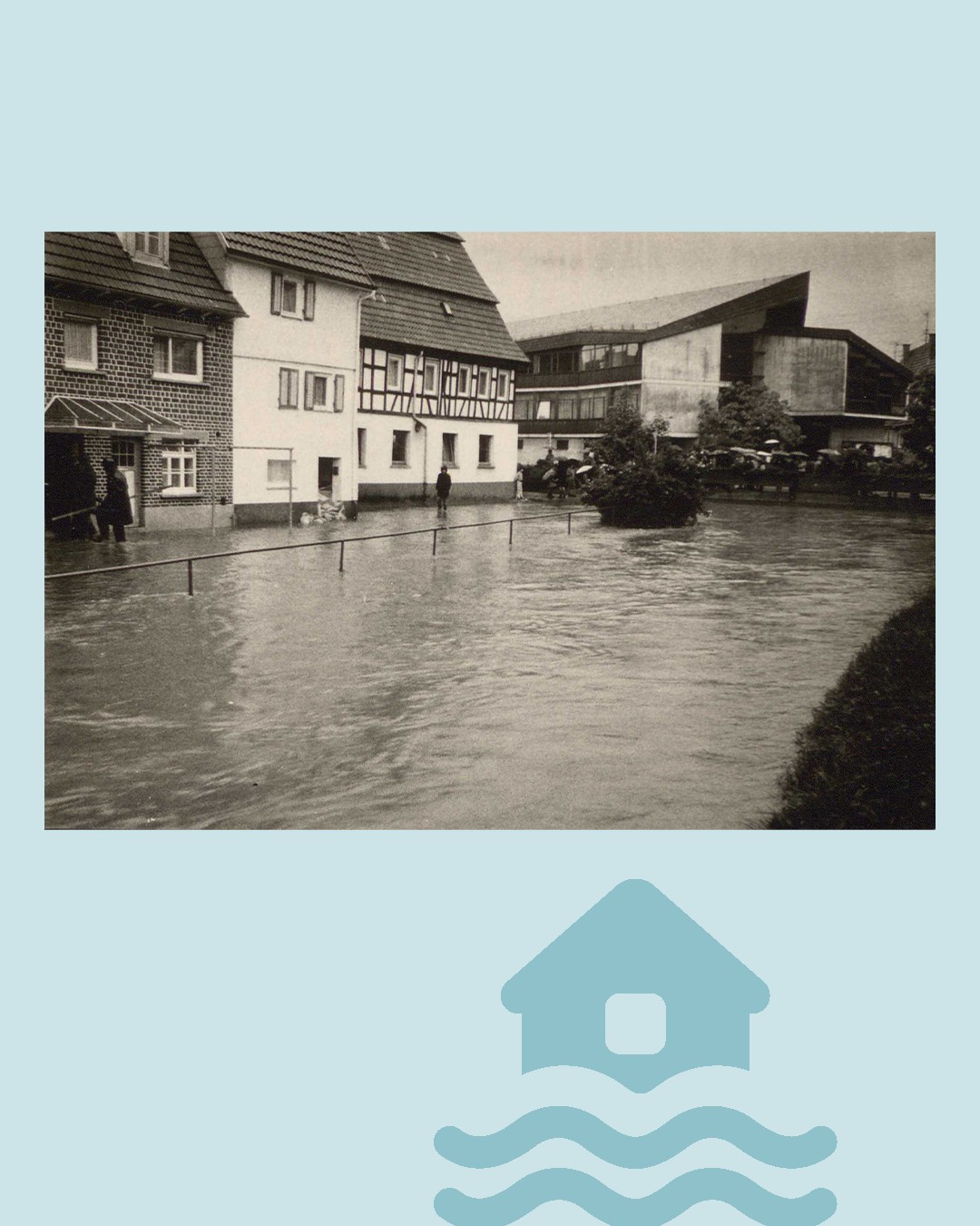 Eine Schwarz-Weiß-Fotografie zeigt eine überflutete Straße, in der mehrere Personen durch das Wasser waten. Links stehen traditionelle Häuser, im Hintergrund ein größeres Gebäude. Am unteren Bildrand ist ein blaues Symbol eines Hauses zu sehen, das von Wellen umspült wird. (KI-Alternativtext)