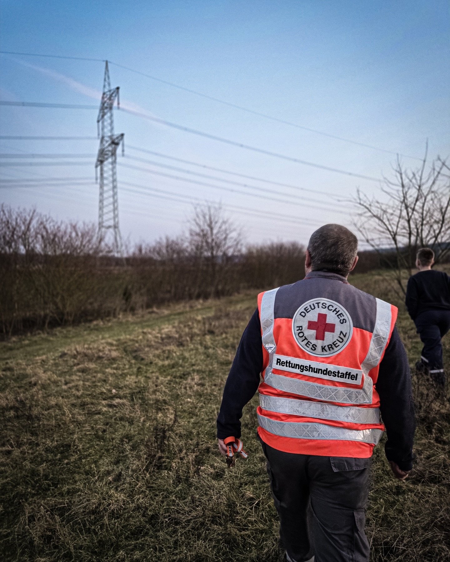 Eine Person von hinten, die eine reflektierende Weste mit dem Logo "Deutsches Rotes Kreuz" und der Aufschrift "Rettungshundestaffel" trägt, geht durch ein grasbewachsenes Feld. In der rechten Hand hält die Person ein orangefarbenes Objekt, während im Hintergrund ein Strommast und eine weitere Person zu sehen sind. (KI-Alternativtext)