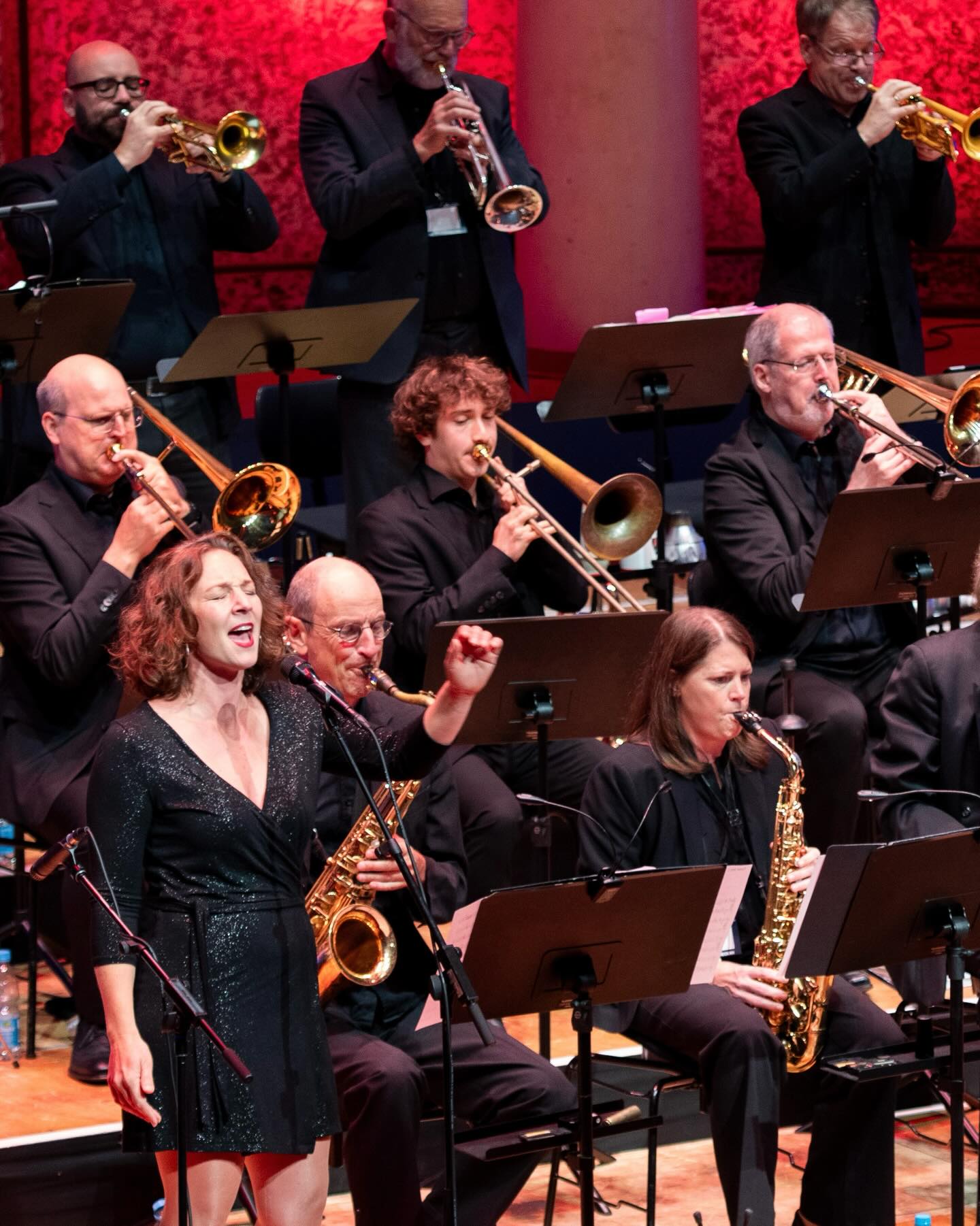 Dieser Alternativtext wurde KI-generiert: Eine Gruppe von Musiker*innen spielt auf einer Bühne. Verschiedene Blasinstrumente wie Trompeten, Posaunen und Saxophone sind zu sehen.