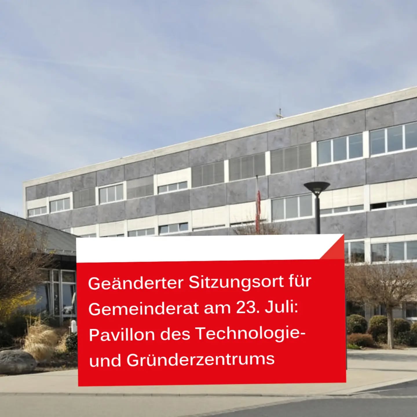 Ein modernes, mehrstöckiges Gebäude mit grauer und weißer Fassade. Davor liegt ein roter Textkasten mit weißer Schrift, der lautet: „Geänderter Sitzungsort für Gemeinderat am 23. Juli: Pavillon des Technologie- und Gründerzentrums“. (KI-Alternativtext)