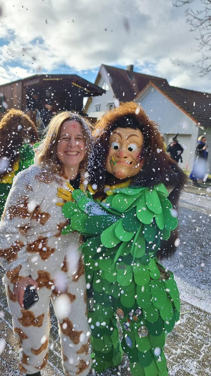 Dieser Alternativtext wurde KI-generiert: Eine Person mit einer anderen in einem farbenfrohen Kostüm umarmt sich im Schnee. Im Hintergrund sind Gebäude und weitere verschwommene Gestalten zu sehen.