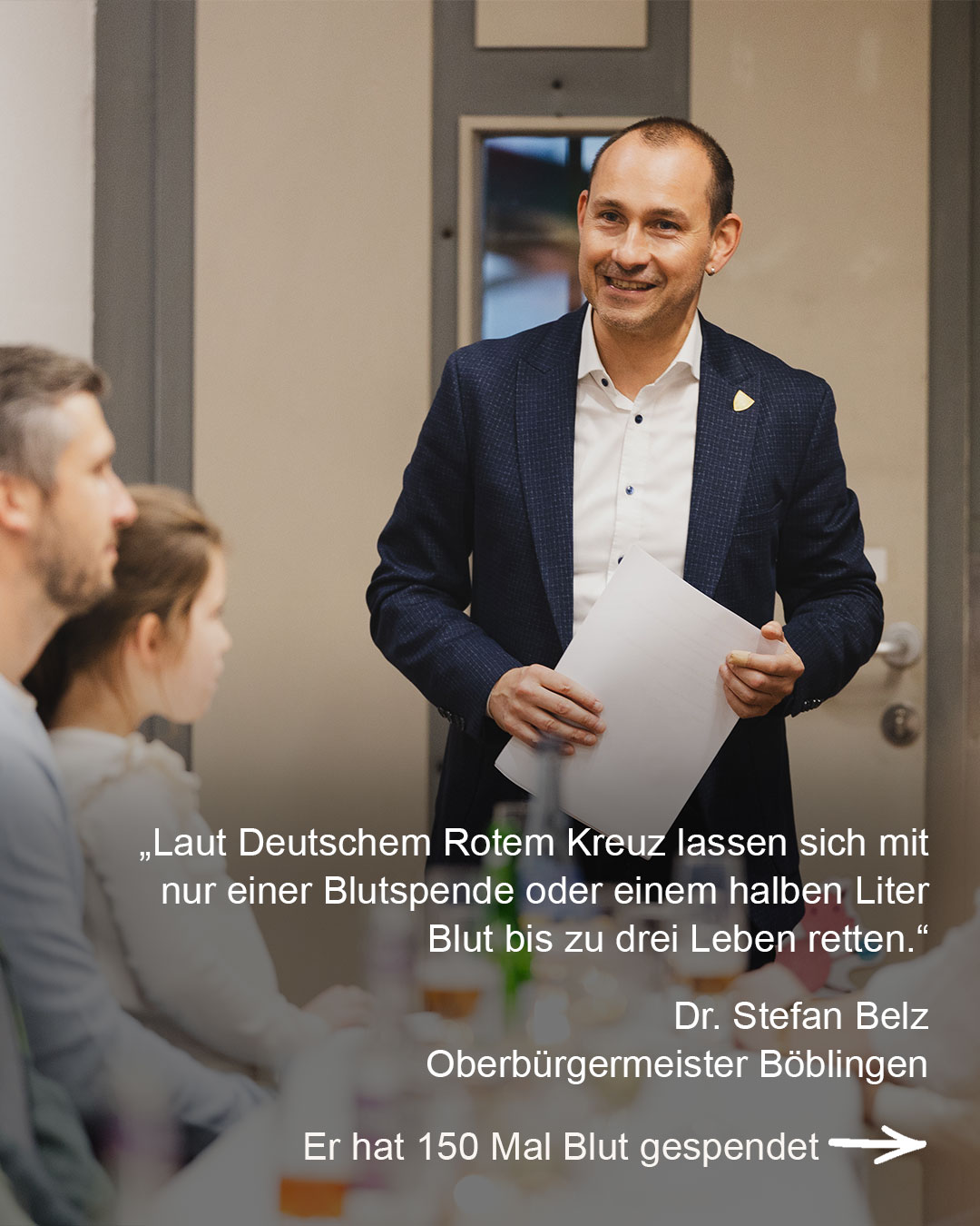 Dieser Alternativtext wurde KI-generiert: Ein Mann in einem dunklen Anzug steht vor einer Tür und spricht. Ein Textzitat über Blutspenden ist im Bild zu sehen.