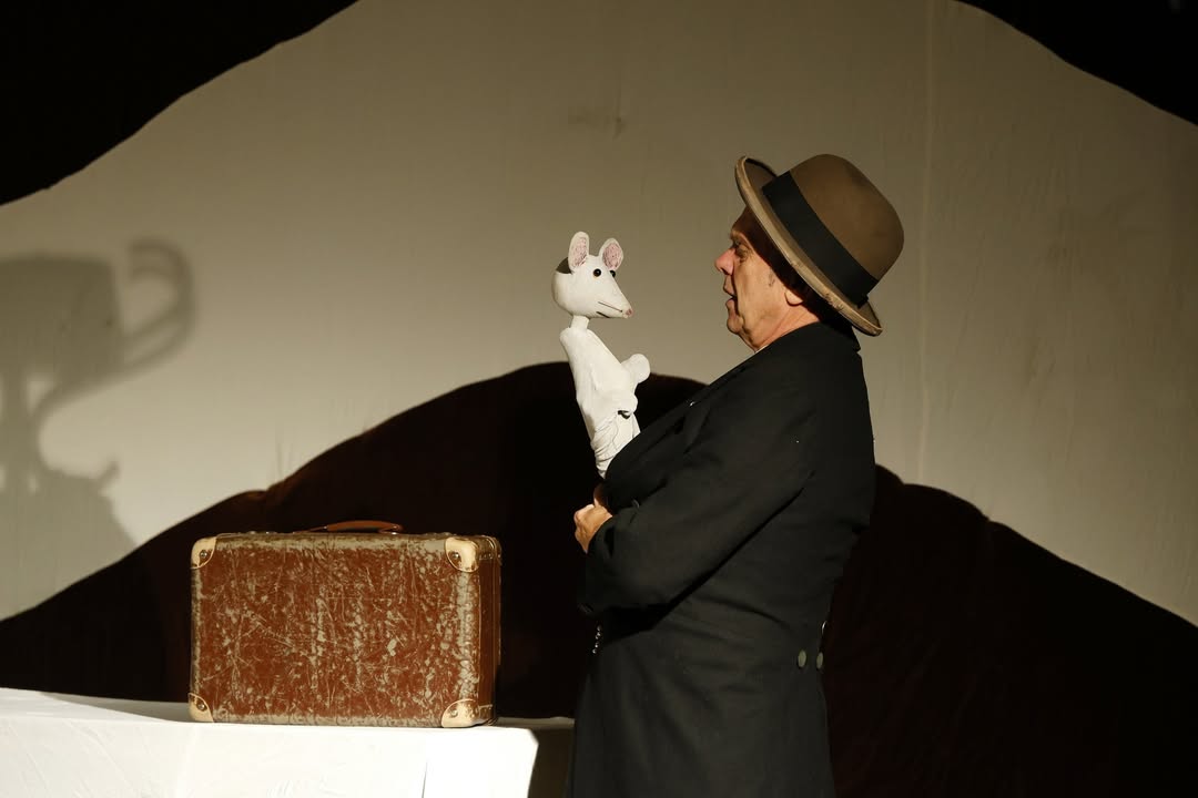 Dieser Alternativtext wurde KI-generiert: Eine Person mit Hut hält eine Stoffmaus in der Hand. Im Hintergrund befindet sich eine Wand mit Schattenspiel und ein Koffer.