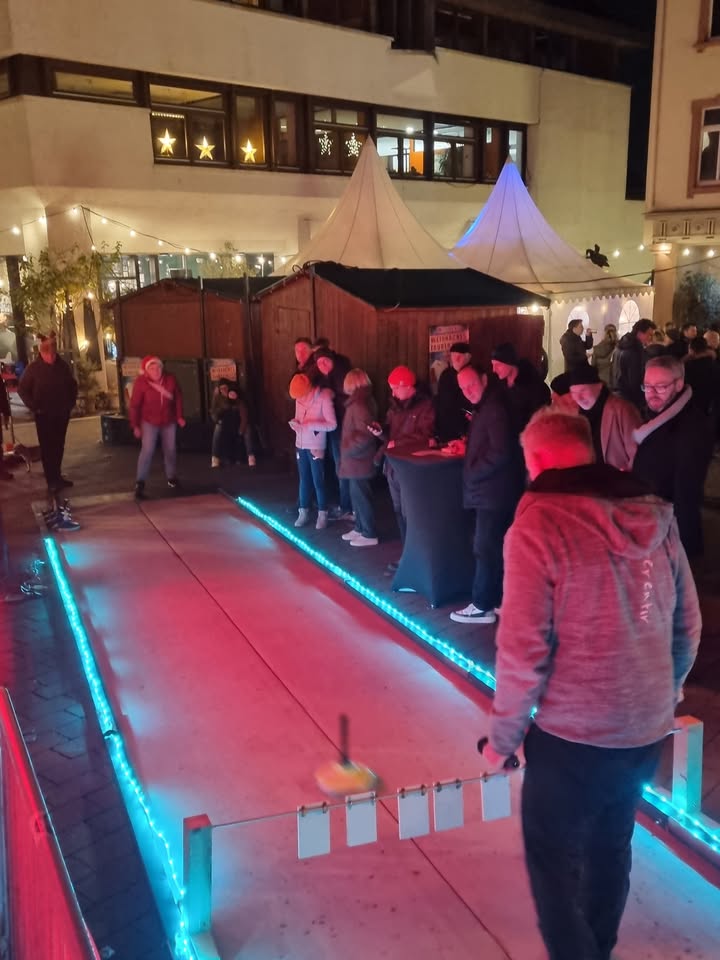 Dieser Alternativtext wurde KI-generiert: Ein belebter Platz mit weihnachtlichen Buden und Besuchern. Lichterketten und festliche Dekorationen sorgen für eine stimmungsvolle Atmosphäre.