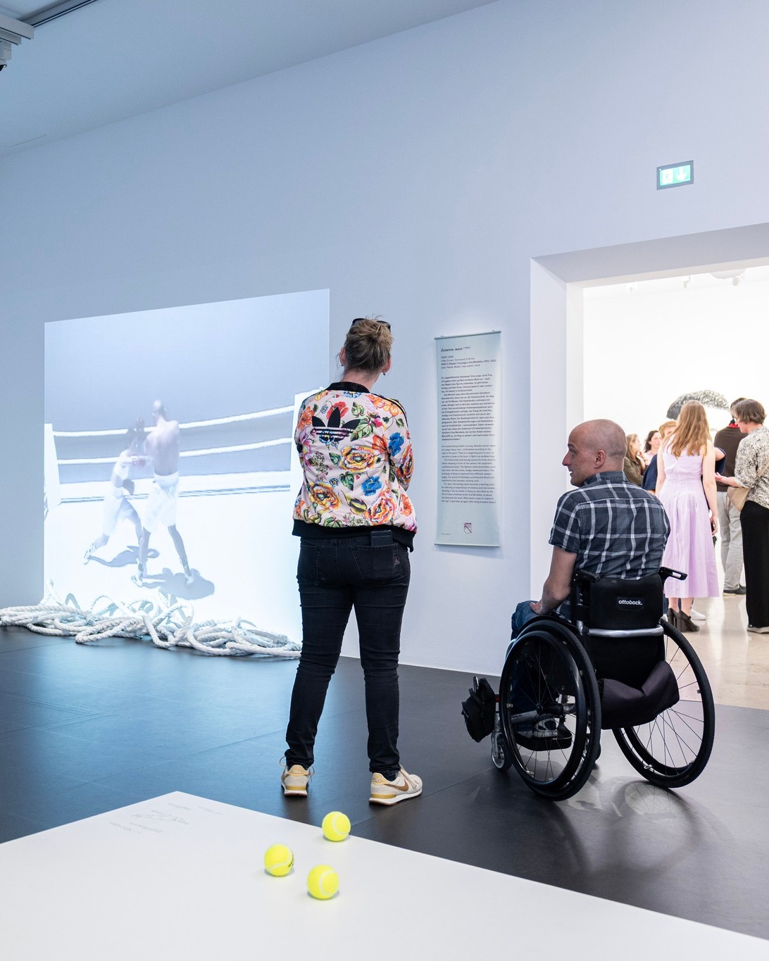 Zwei Personen betrachten in einer Ausstellung eine Wandprojektion, die zwei Boxer in einem Ring zeigt. Eine Person steht in einer bunten Jacke, während die andere Person im Rollstuhl sitzt. Im Vordergrund liegen drei gelbe Tennisbälle auf einem weißen Tisch, am Boden liegen dicke Seile. (KI-Alternativtext)