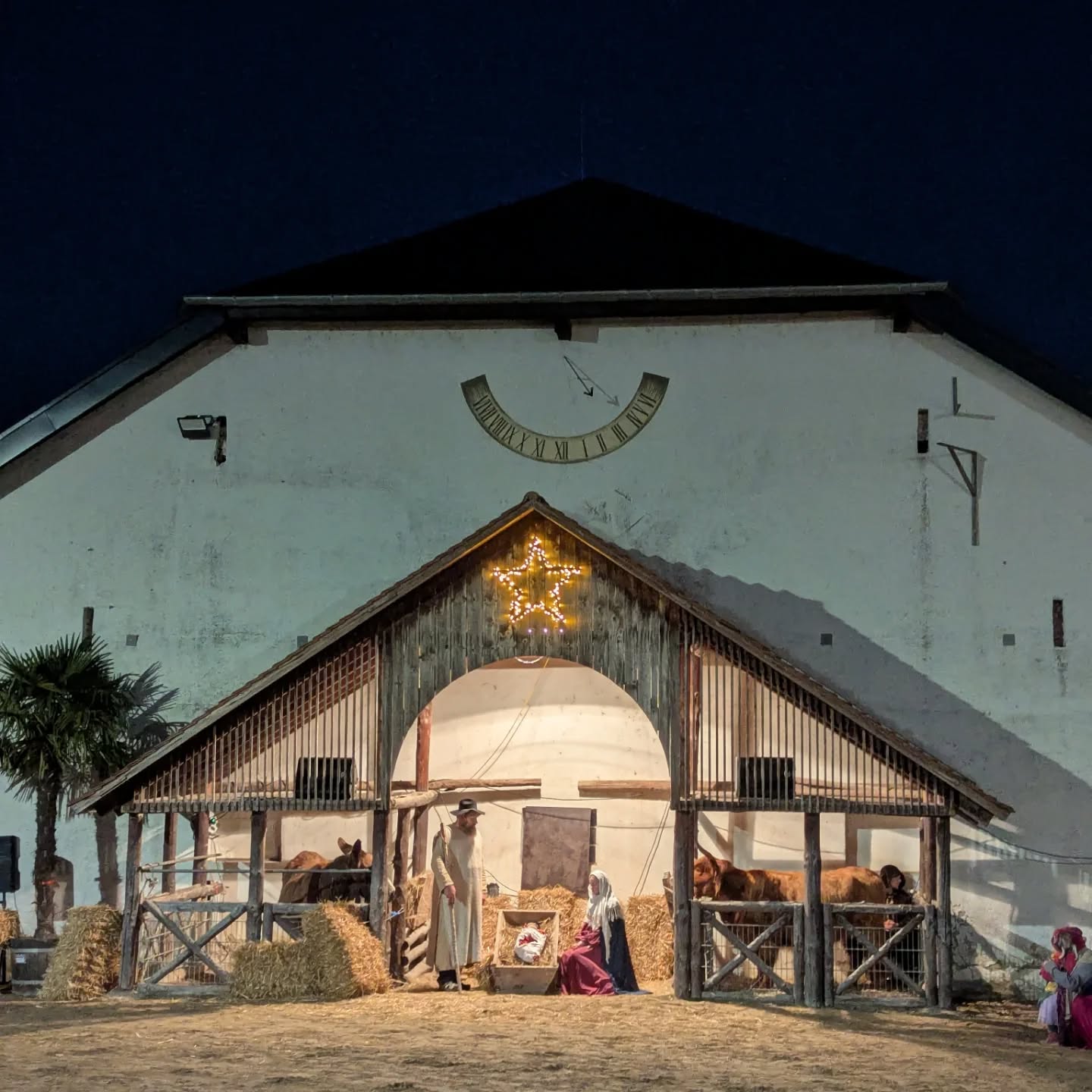 Dieser Alternativtext wurde KI-generiert: Ein hell erleuchteter Stall mit einer Darstellung einer Krippenszene im Freien. Über dem Stall befindet sich eine große Uhr.