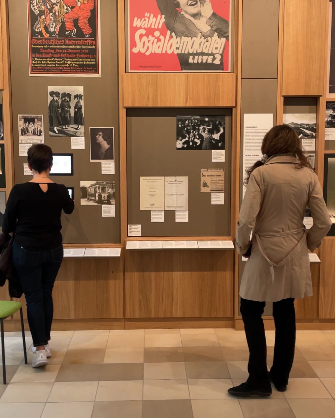 Zwei Personen betrachten aufmerksam Ausstellungsstücke in einem Museum. Die Exponate umfassen Plakate, Fotografien und Dokumente.