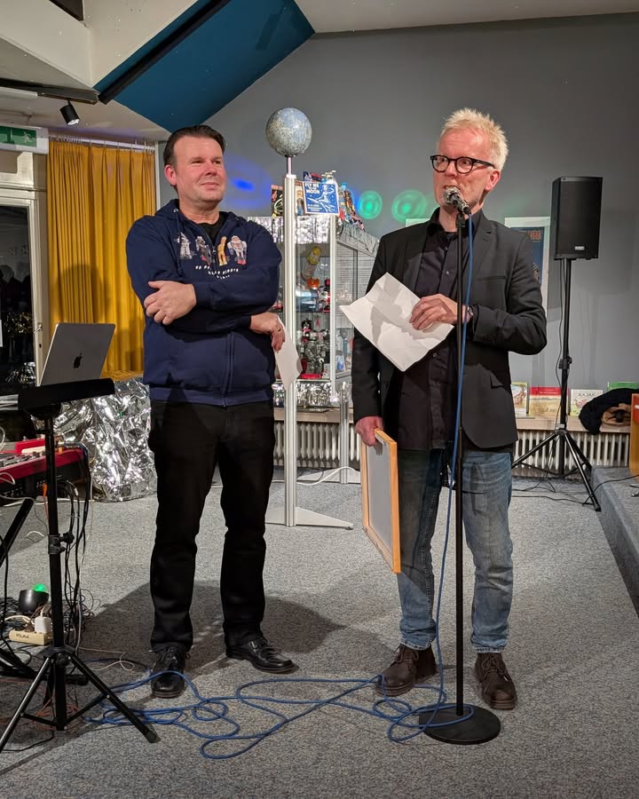 Dieser Alternativtext wurde KI-generiert: Zwei Personen stehen auf einer Bühne und sprechen in Mikrofone. Im Hintergrund sind ein beleuchteter Raum und technisches Equipment zu sehen.