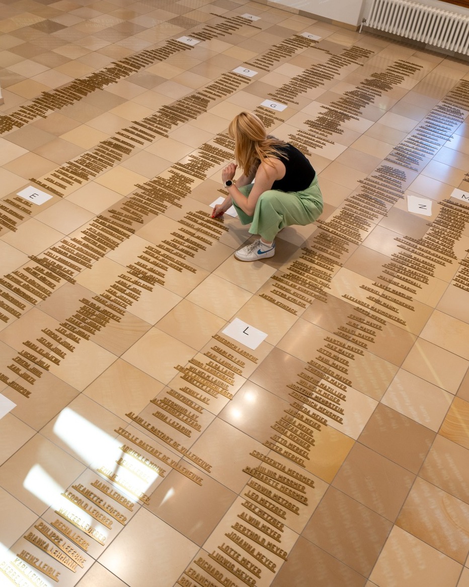 Eine Person hockt auf einem hellen Holzboden und betrachtet eine Installation aus zahlreichen Namen, die in alphabetischer Reihenfolge auf dem Boden angebracht sind. Weiße Zettel mit Buchstaben wie A, E, L und Z markieren die Spalten. Die Person berührt mit der Hand einen der Namen. (KI-Alternativtext)