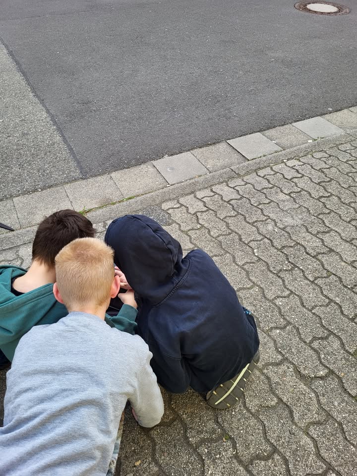 Dieser Alternativtext wurde KI-generiert: Zwei Personen sitzen auf einem Bordstein und schauen auf etwas vor sich. Der Blickwinkel zeigt die Hinterköpfe und Teile der Oberkörper der Personen.