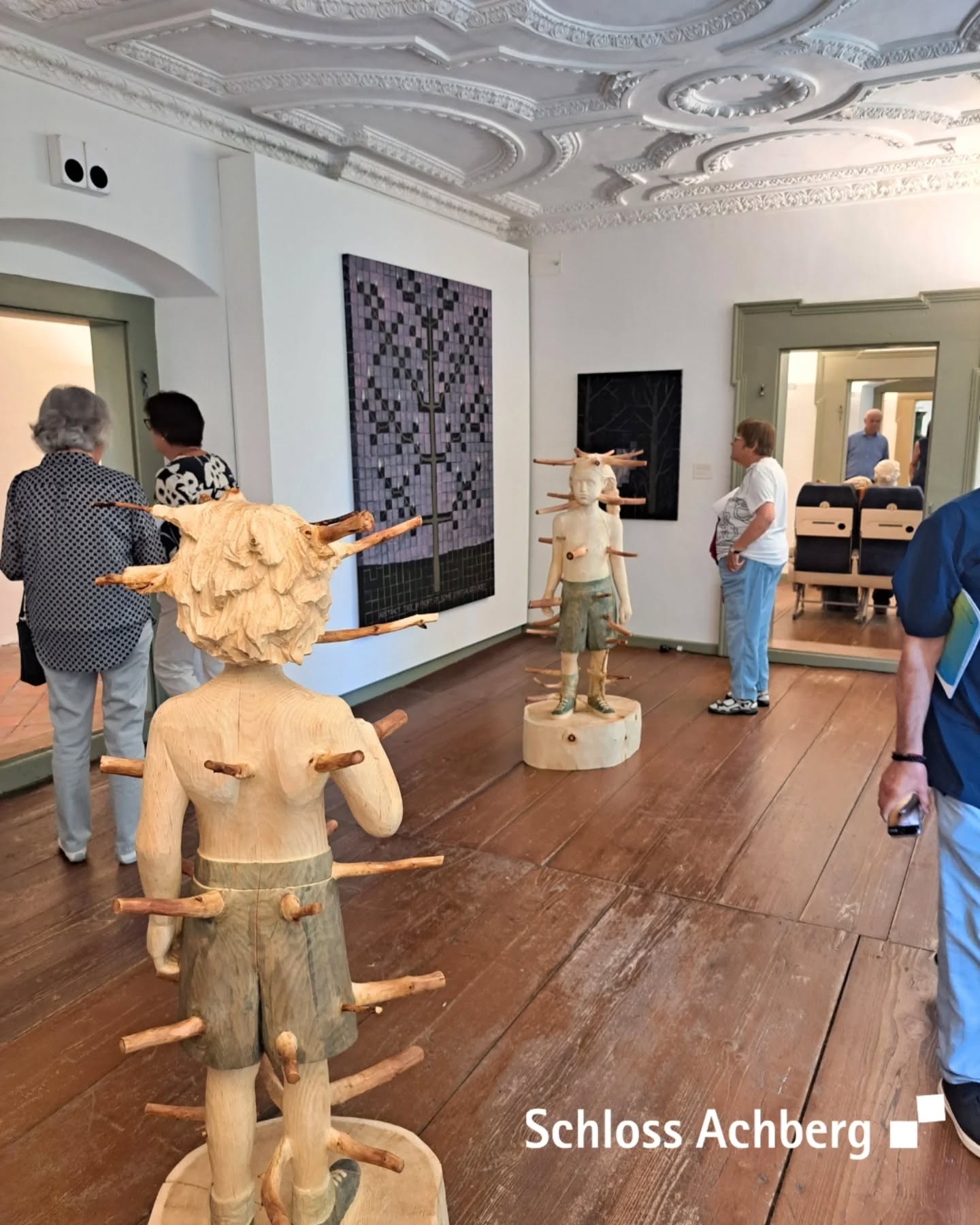 In einem Raum befinden sich mehrere Personen und Holzstatuen mit Ästen darin sowie Bilder an den Wänden. Der Boden besteht aus breiten Holzdielen, und die Decke ist mit einem kunstvollen Muster verziert.