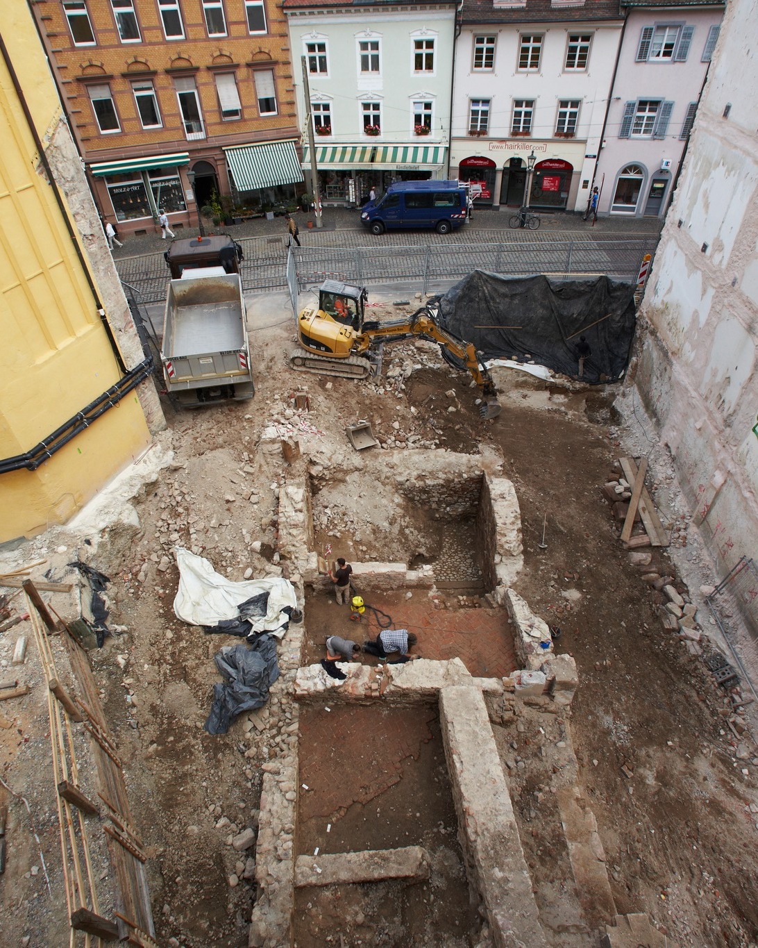 Dieser Alternativtext wurde KI-generiert: Ein Blick von oben zeigt eine Baustelle in einer Stadt. Es werden Ausgrabungen durchgeführt und es sind mehrere Fahrzeuge und Personen zu sehen.