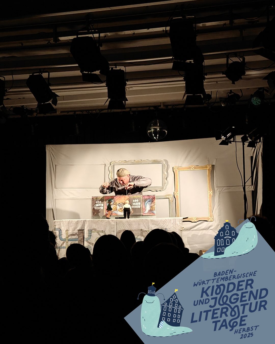 Dieser Alternativtext wurde KI-generiert: Eine Bühne mit einer Person, die an einem Tisch sitzt. Im Hintergrund eine Werbetafel und eine Silhouette von Zuschauern in einem dunklen Raum.